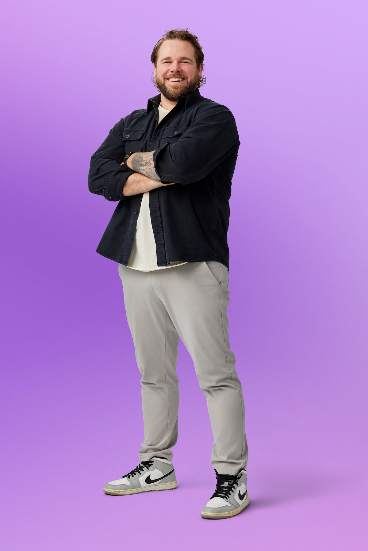 Jordan standing with arms crossed, wearing a dark jacket, light shirt, light pants, and sneakers, on a purple gradient studio background.