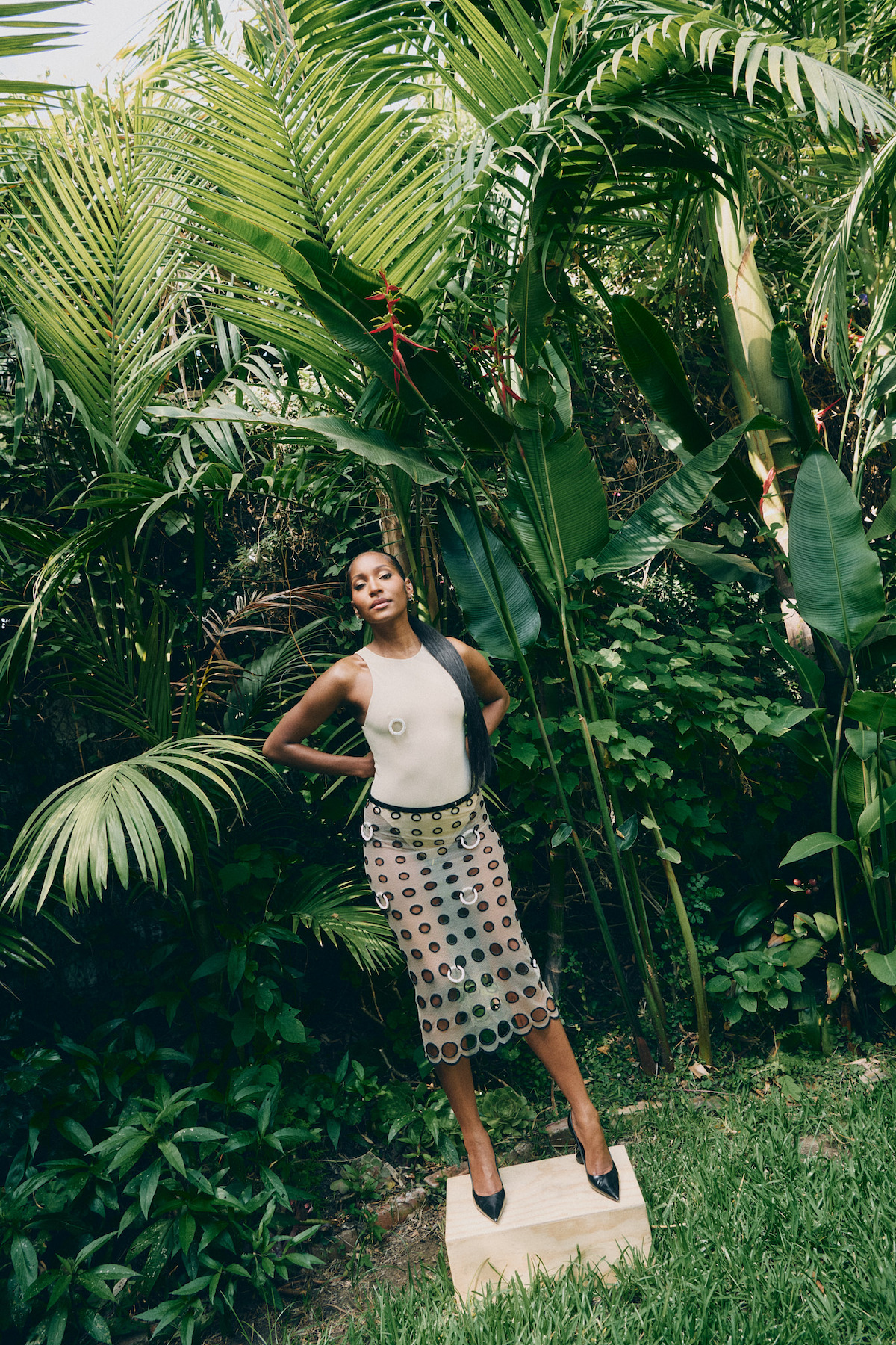 A woman poses outdoors in a lush, tropical garden, standing confidently on a stone surrounded by green plants and palm leaves, wearing a white top, patterned skirt, and black heels.