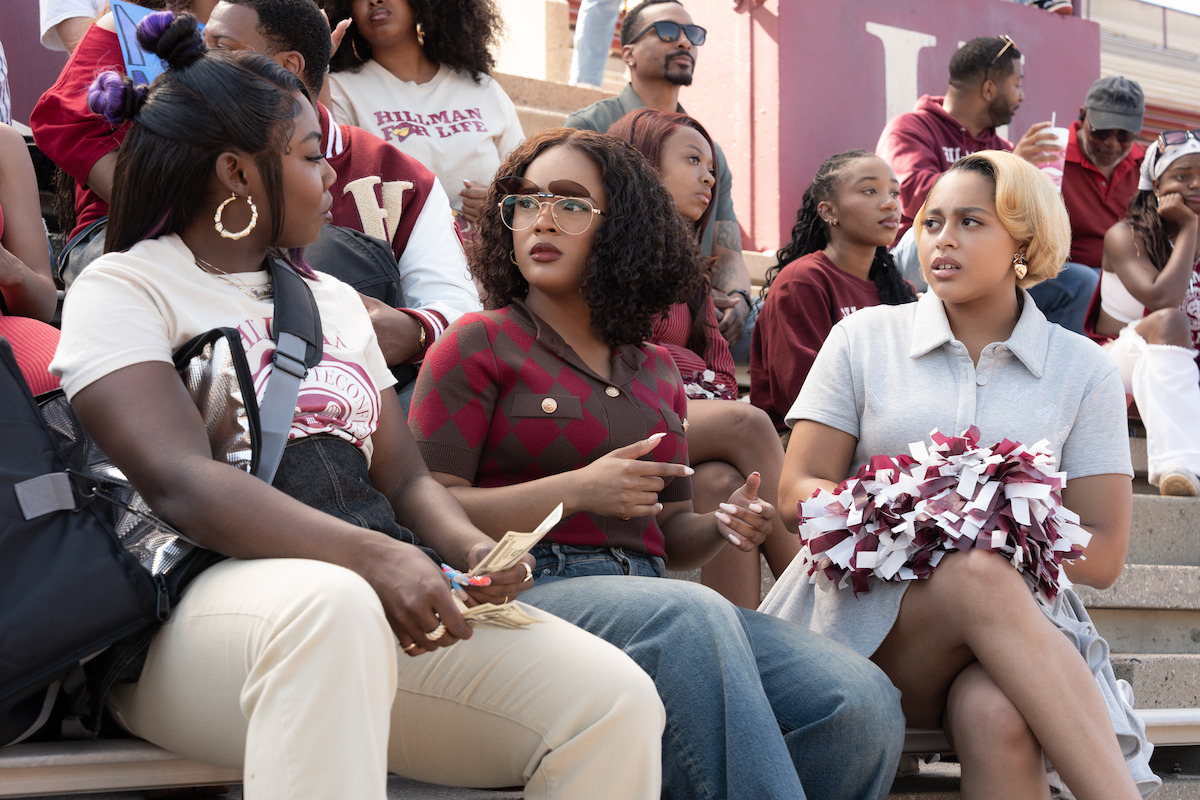 Three young women sit in stadium bleachers during a school event, surrounded by people in maroon and white, some cheering and holding pom-poms, creating a lively, social atmosphere.