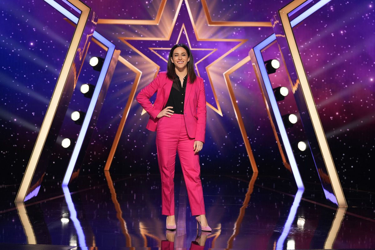 Janine Harouni in a bright pink suit stands confidently on a shiny stage with a large illuminated star and colorful lights in the background, creating a vibrant, energetic show atmosphere.