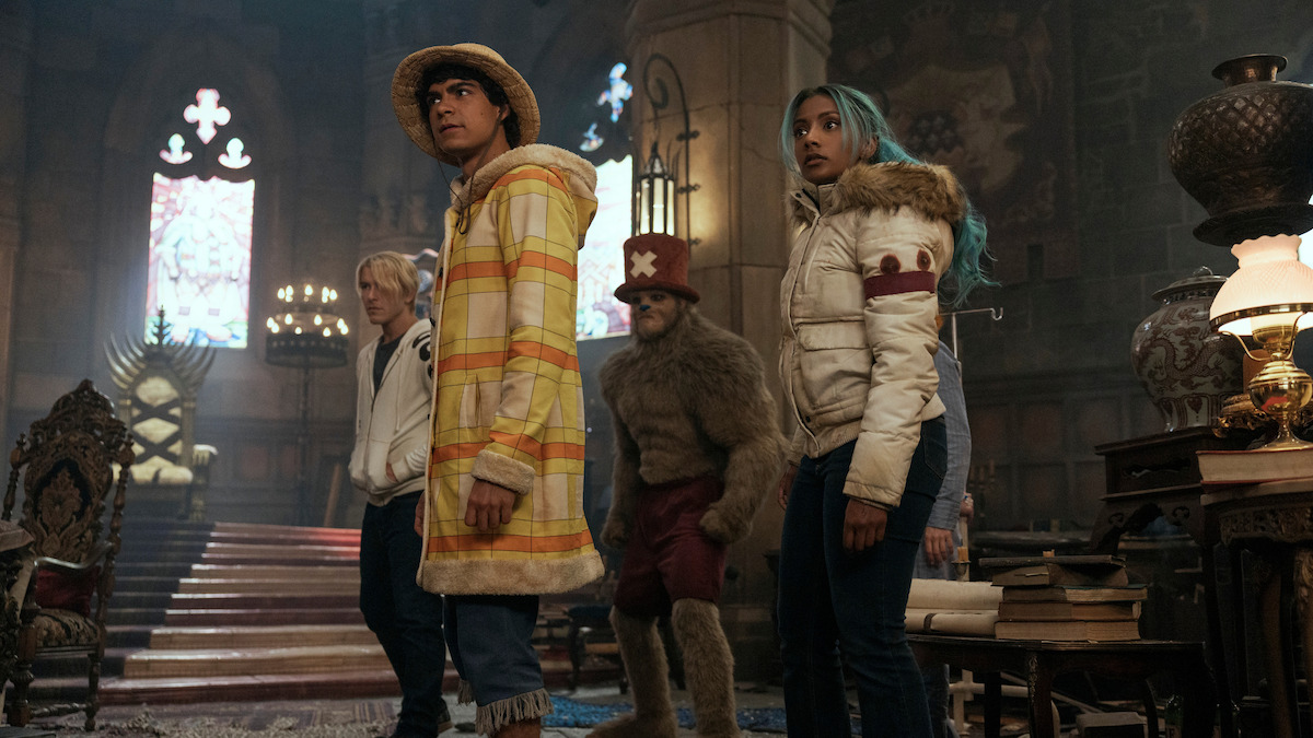 Four people in colorful clothing stand alert in a grand, dimly lit room with gothic architecture, large candles, ornate chairs, books, and stained glass windows, suggesting a dramatic or fantasy setting.