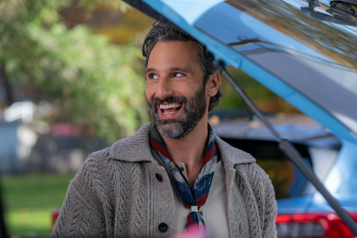 Smiling man with gray hair and beard stands outdoors by an open car trunk, wearing a knitted sweater and scarf, with green trees and a blurred background suggesting a park or suburban setting.