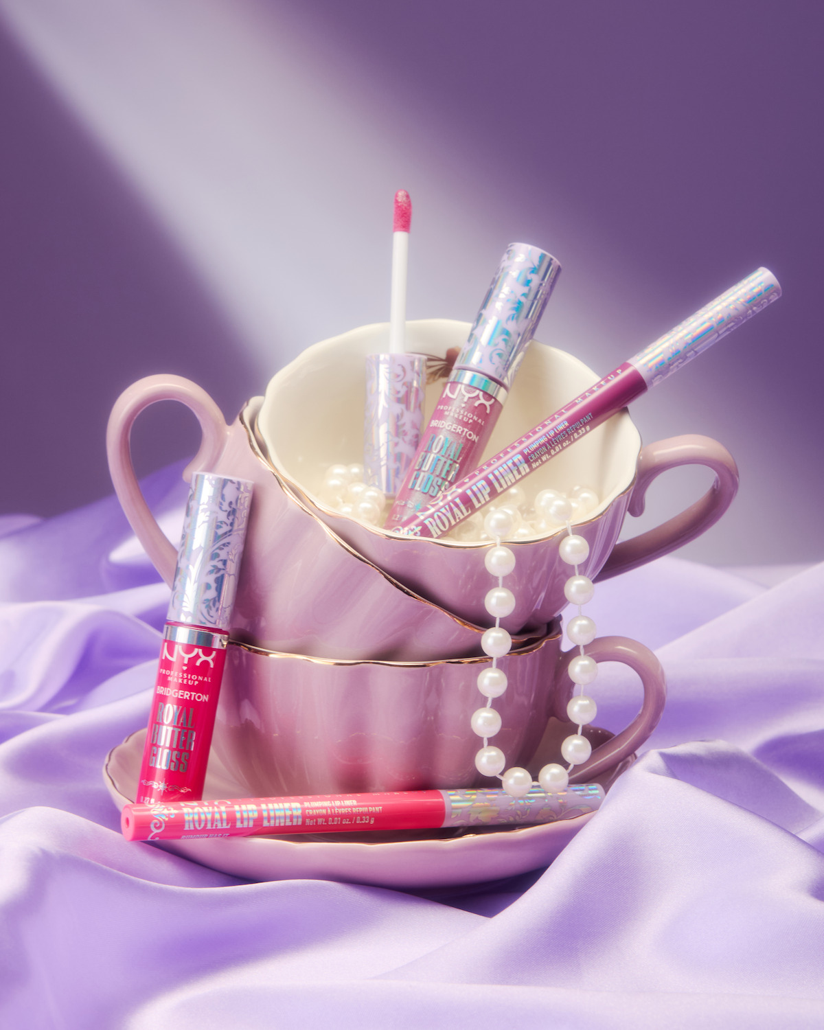 Pink tea cups stacked with NYX makeup products, a pearl necklace, and satin fabric arranged in soft, pastel lighting for a whimsical, elegant display.