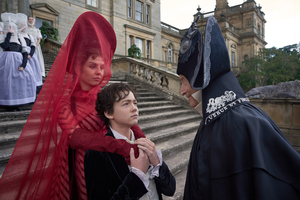 Three people in elaborate historical costumes stand on stone steps of a grand building; the mood is tense and dramatic, with one in red clutching another, and a third in black facing them closely. Others in period attire are in the background.