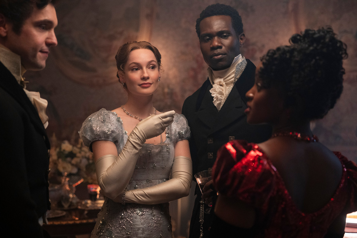 Four people in elegant Regency-era costumes converse in a dimly lit, ornately decorated room, suggesting a historical or period drama setting, with one woman in a silver gown and another in red.