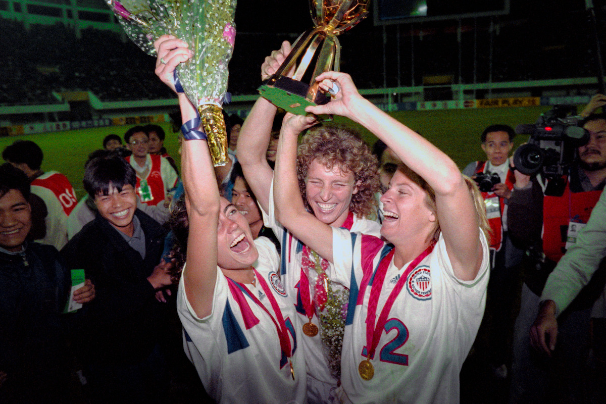 Three jubilant female soccer players hold a trophy, gold medals, and flowers, celebrating victory on a stadium field at night with cheering teammates and media in the background.