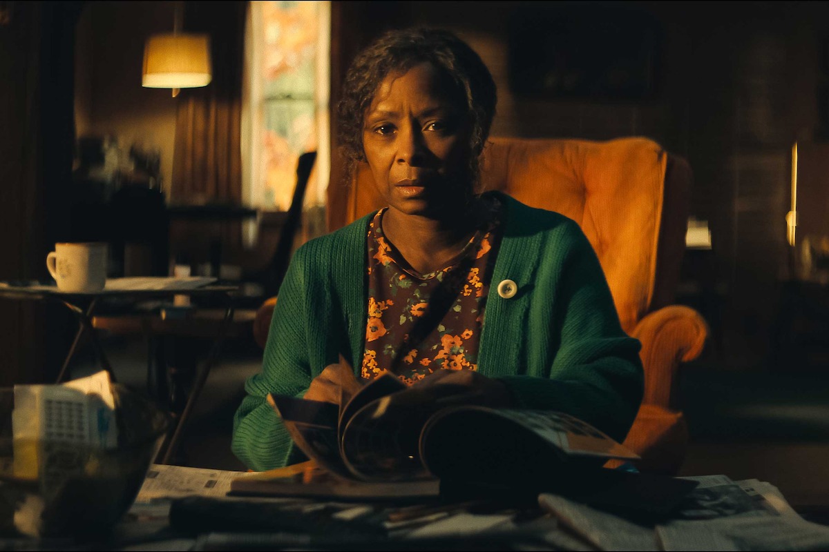 Older woman in a green cardigan sits in an orange chair at a cluttered table with magazines and papers, in a warmly lit living room with vintage decor and a lamp in the background, looking intently forward.