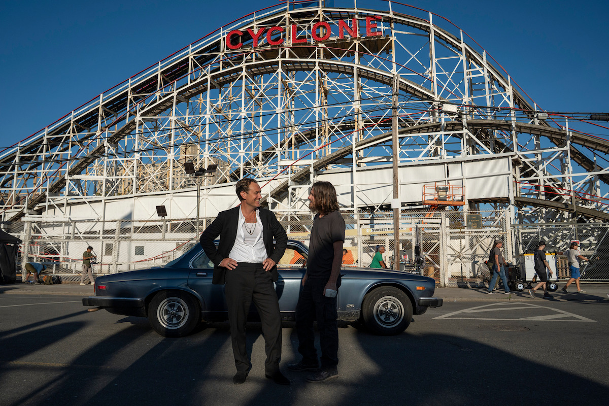 Two people stand in front of a vintage car at an amusement park, with a large wooden roller coaster labeled "Cyclone" in the background on a sunny day.