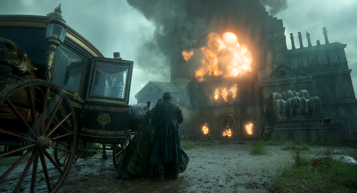 Two people in period clothing stand near a carriage in heavy rain, watching a large, ornate building engulfed in flames and smoke, creating a dramatic and tense atmosphere.