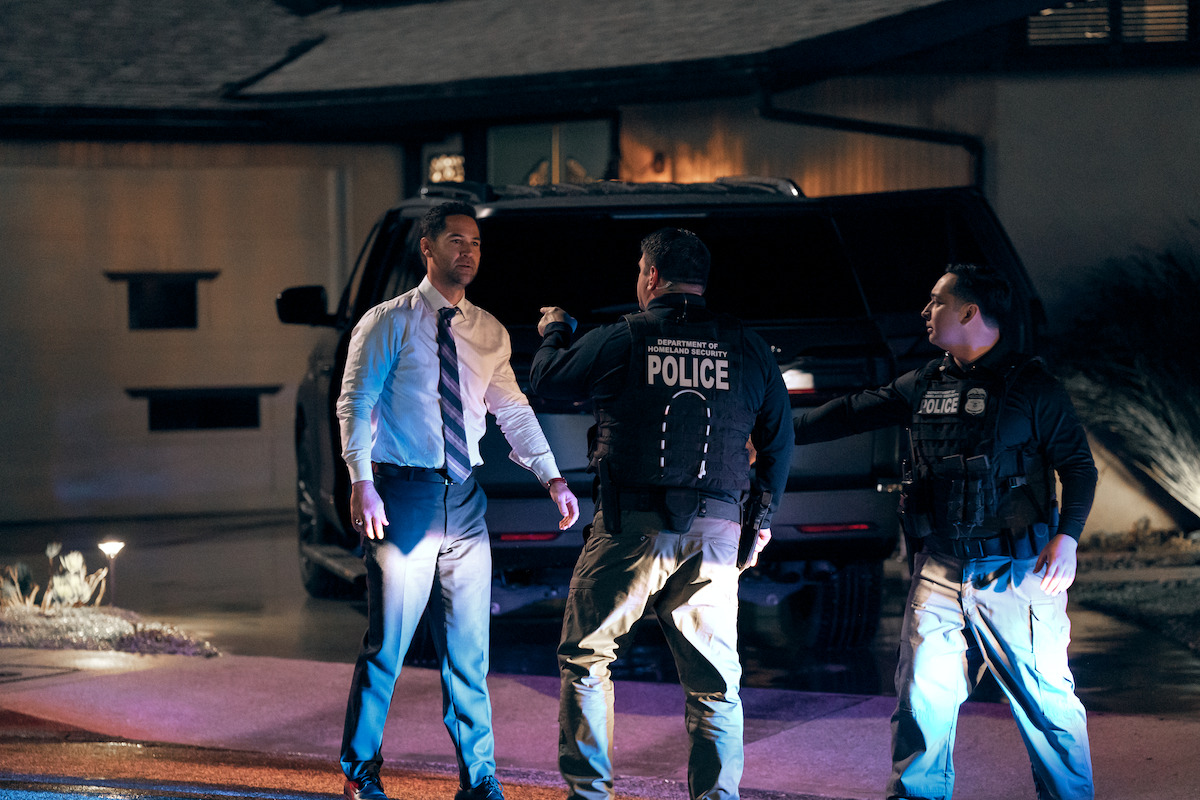 Three police officers stand in front of a black SUV parked outside a house at night, illuminated by artificial lighting, one in a shirt and tie and two in tactical gear marked "POLICE".