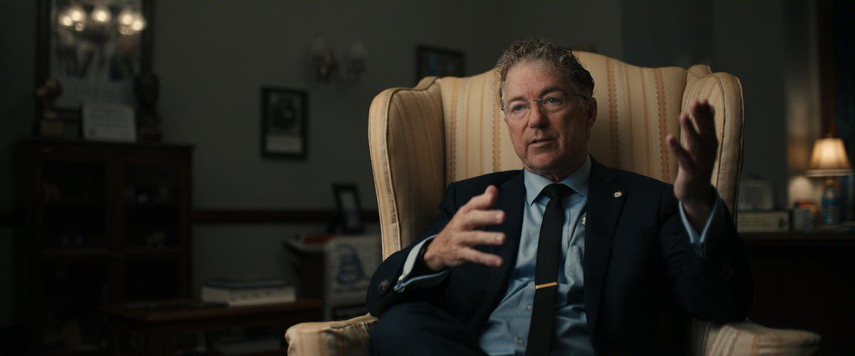 Man in a suit sits in a striped armchair in a dimly lit, formal office or study, gesturing with both hands while speaking. Books, framed pictures, and lamps fill the background, creating a professional atmosphere.