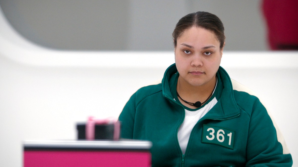 A woman in a green tracksuit with the number 361 sits indoors at a table in a bright, modern setting, looking serious and focused, evoking a tense, contemplative mood.