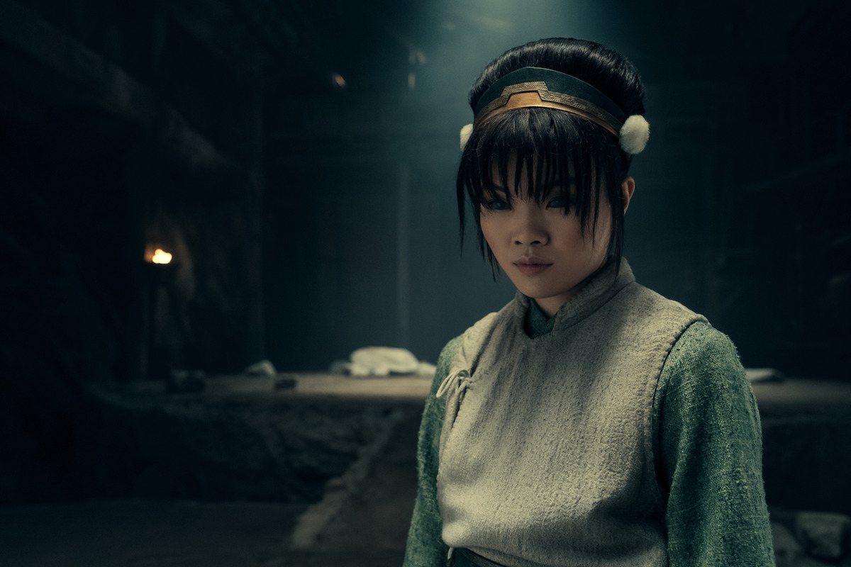 Miyako as Toph with dark hair and traditional attire stands in a dimly lit, stone-walled underground setting, looking downward thoughtfully. Warm torchlight glows in the shadowy, cavernous background.