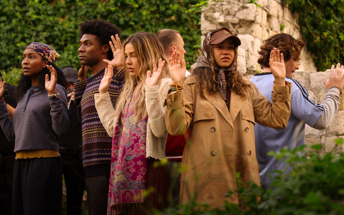 A group of people standing outdoors near a stone wall, raising their hands in surrender, surrounded by greenery.