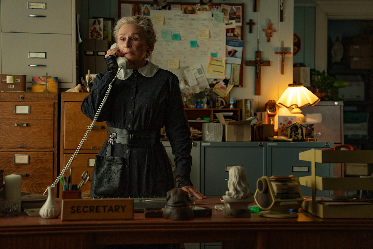 Older woman in vintage dress stands behind cluttered secretary desk, talking on a corded phone in a dimly lit, busy office with files, papers, and religious decorations, creating a serious, focused mood.