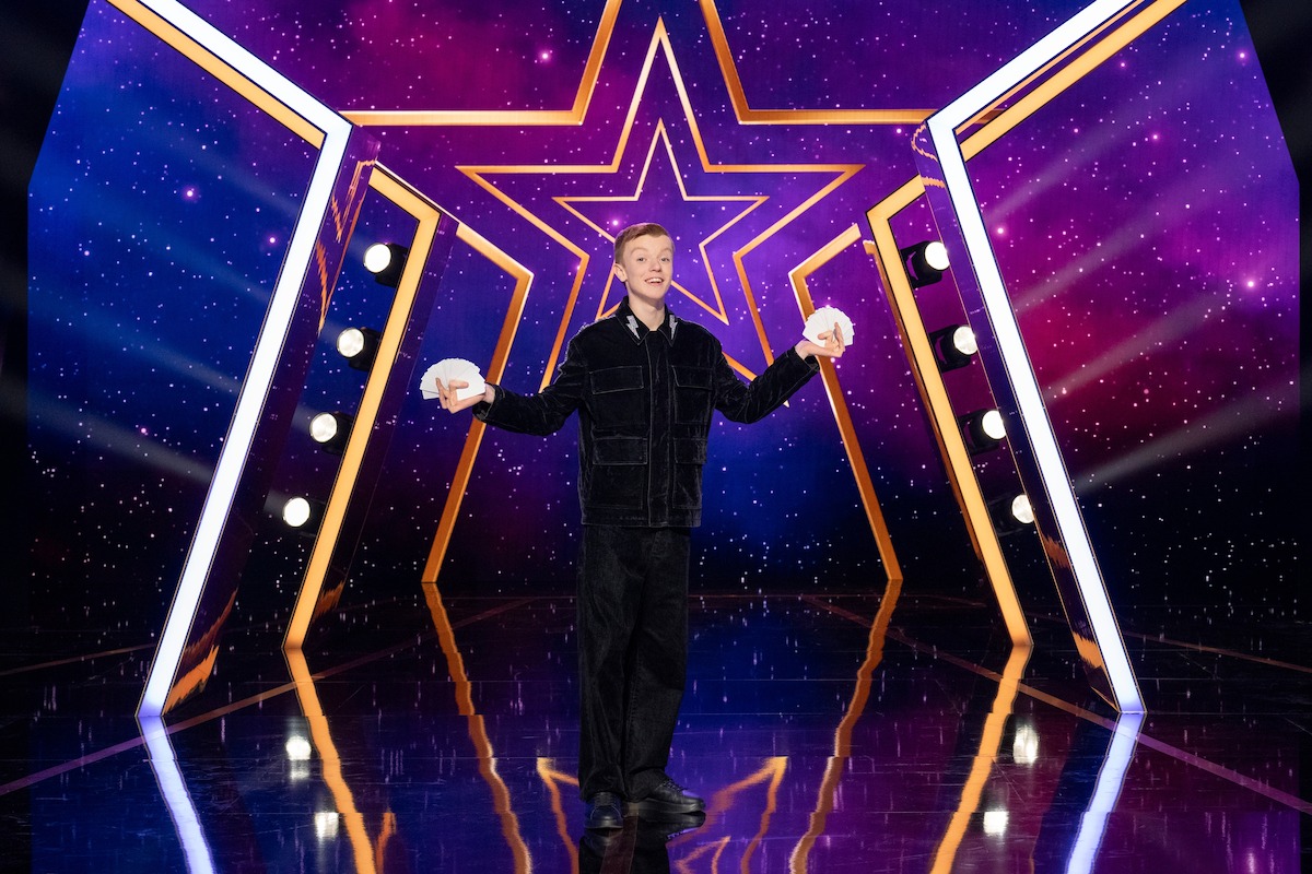 Harry Merlin Piper actuando en un escenario brillantemente iluminado con un fondo de estrellas, sosteniendo cartas en ambas manos, rodeado de focos y un fondo púrpura de temática cósmica.