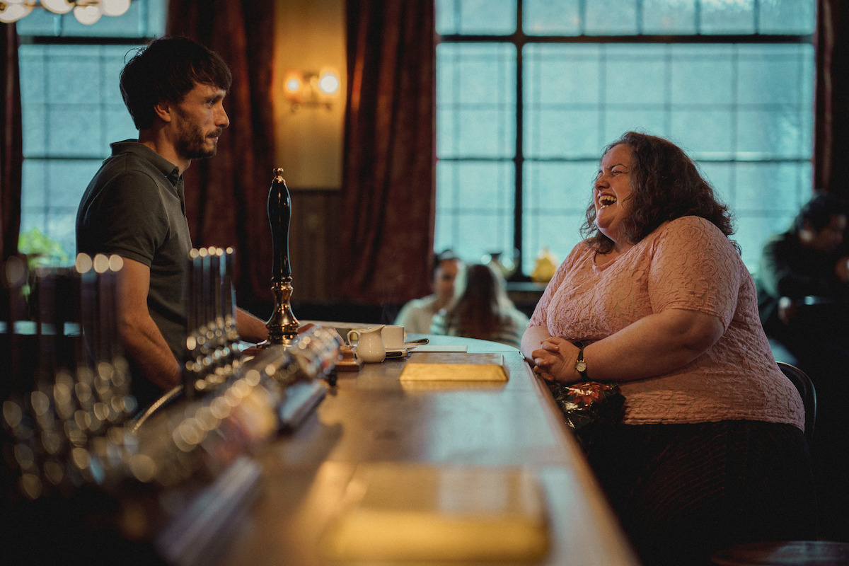 Richard Gadd as Donny Dunn stands behind a bar while Jessica Gunning as Martha sits at the other side laughing in ‘Baby Reindeer’