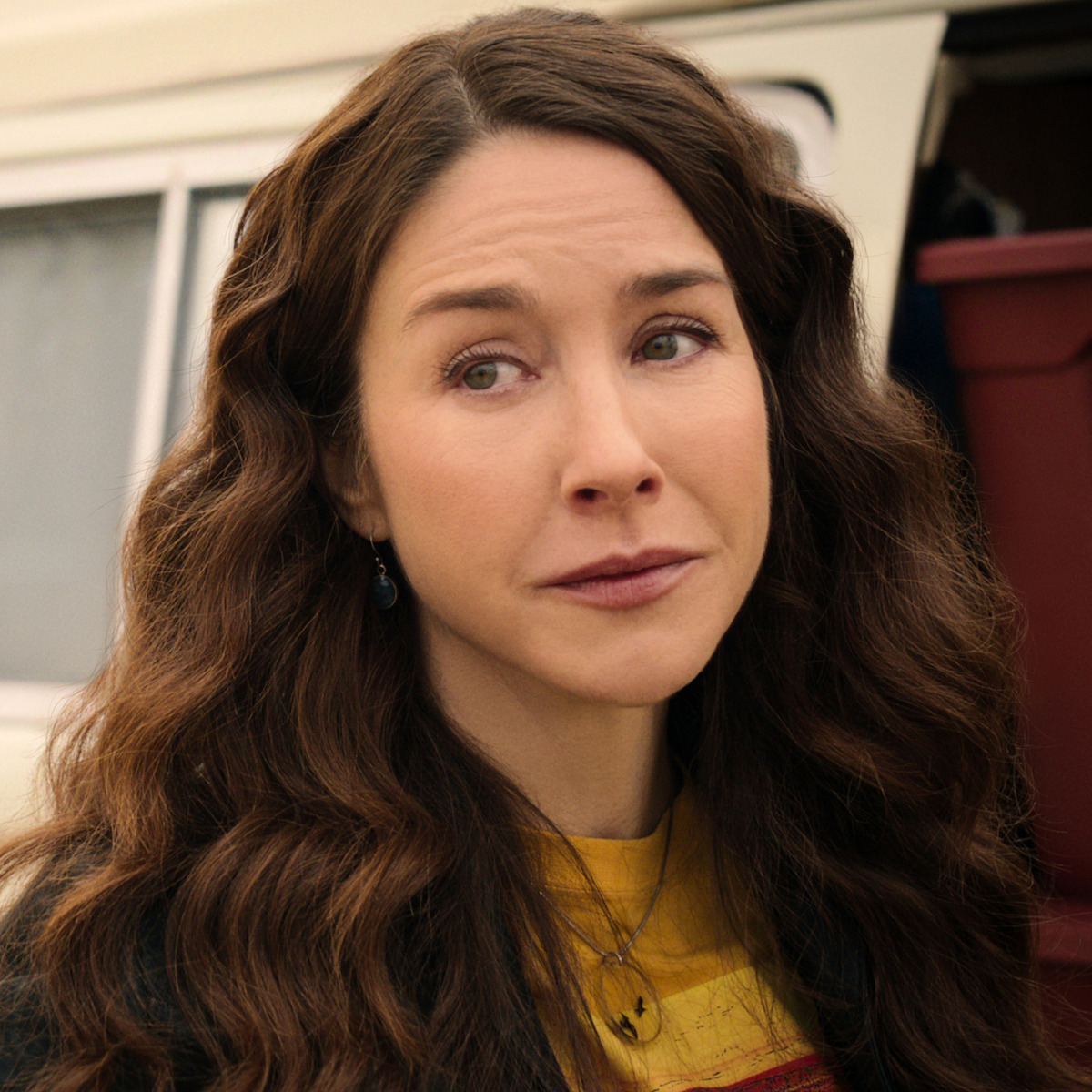 Woman with long wavy brown hair, wearing a yellow shirt and necklace, stands outdoors next to a vehicle with a red storage bin in the background, looking pensive or emotional.