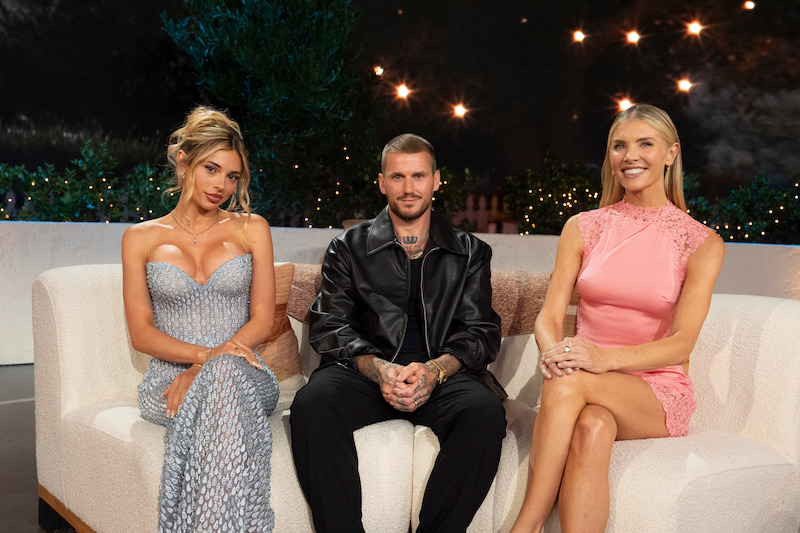 Three people sitting on a couch; two women in elegant dresses and one man in a leather jacket, in a formal, studio-like setting with decorative plants and string lights in the background.
