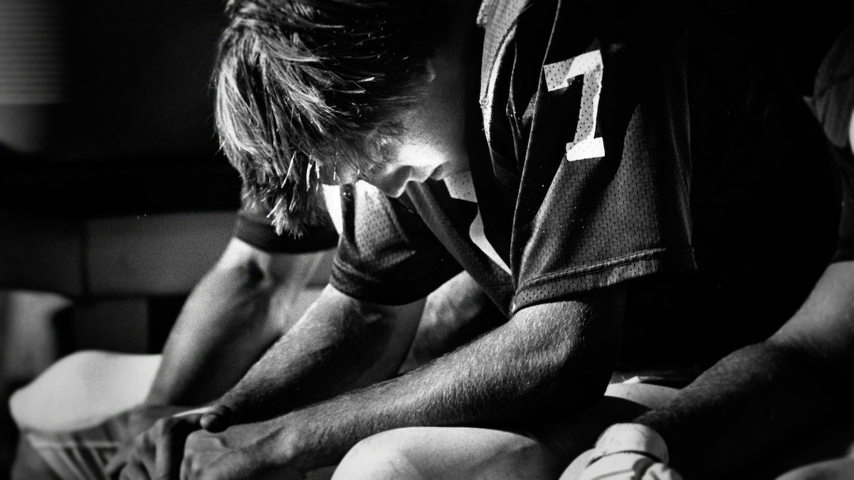 John Elway in uniform sits on a bench in a dimly lit locker room, head bowed and hands clasped, appearing deep in thought or reflection before or after a game.