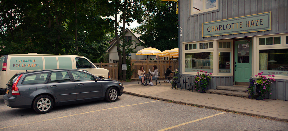 People sit at outdoor tables with yellow umbrellas in front of a bakery café called Charlotte Haze. Two cars are parked nearby, one labeled "Patisserie Boulangerie." Trees and a home are visible in the background.