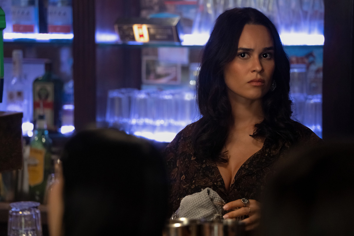 Woman with dark hair behind a bar, holding a towel and glass, looking serious. Shelves with bottles and glasses are visible behind her in a dimly lit bar setting. Other people are slightly out of focus in the foreground.