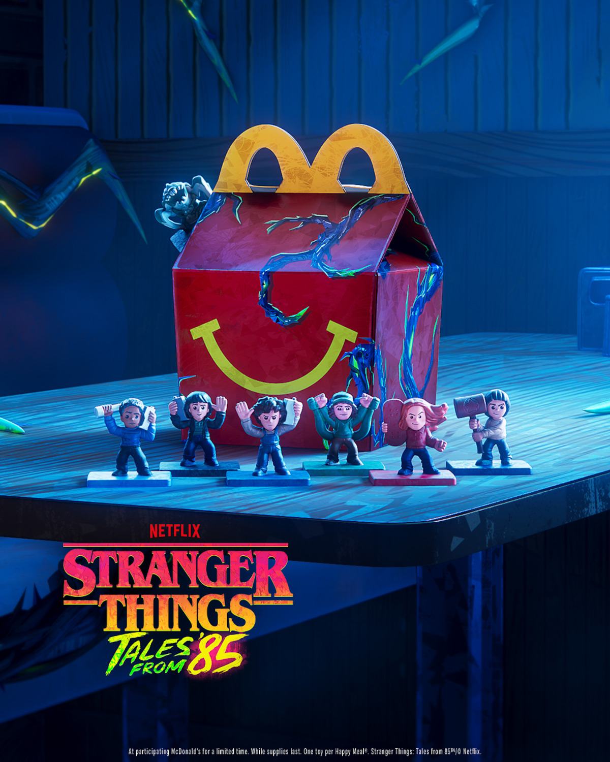 A cracked McDonald's Happy Meal box sits on a table surrounded by Stranger Things character toys in a dimly lit room with glowing cracks, promoting Netflix's Stranger Things: Tales from 85.