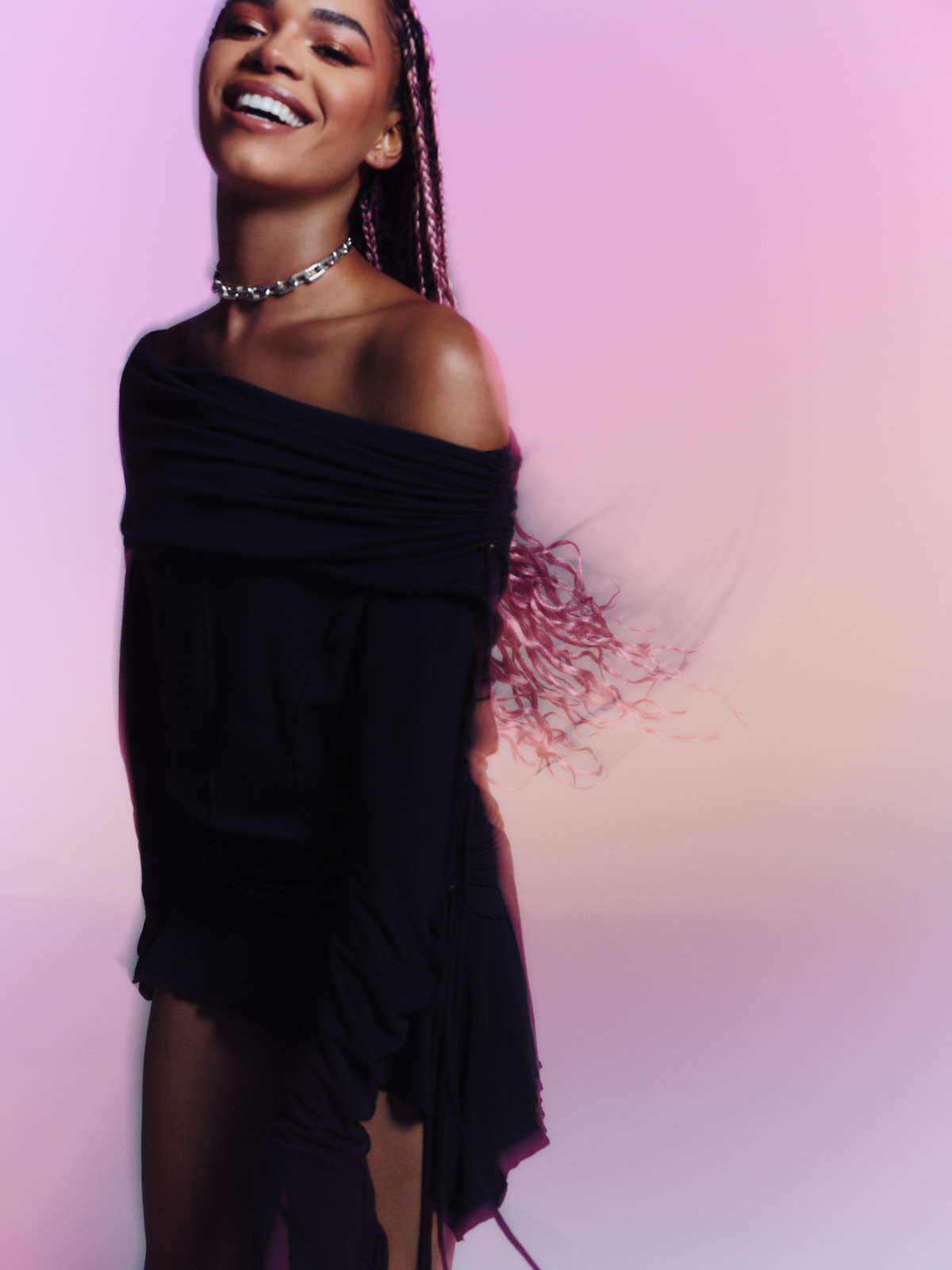 Smiling woman with braided hair, wearing a black off-shoulder outfit and silver choker, standing in front of a pink and purple gradient background with motion blur in her hair.