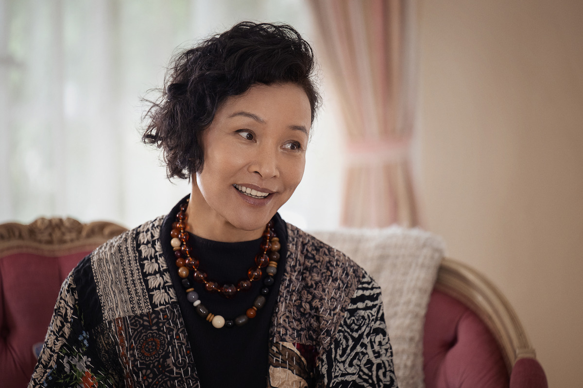 Joan Chen as Janice, with short curly hair, wearing layered necklaces and a patterned jacket, sits on a vintage pink sofa in a softly lit, elegant room with sheer curtains and pastel drapes in the background.
