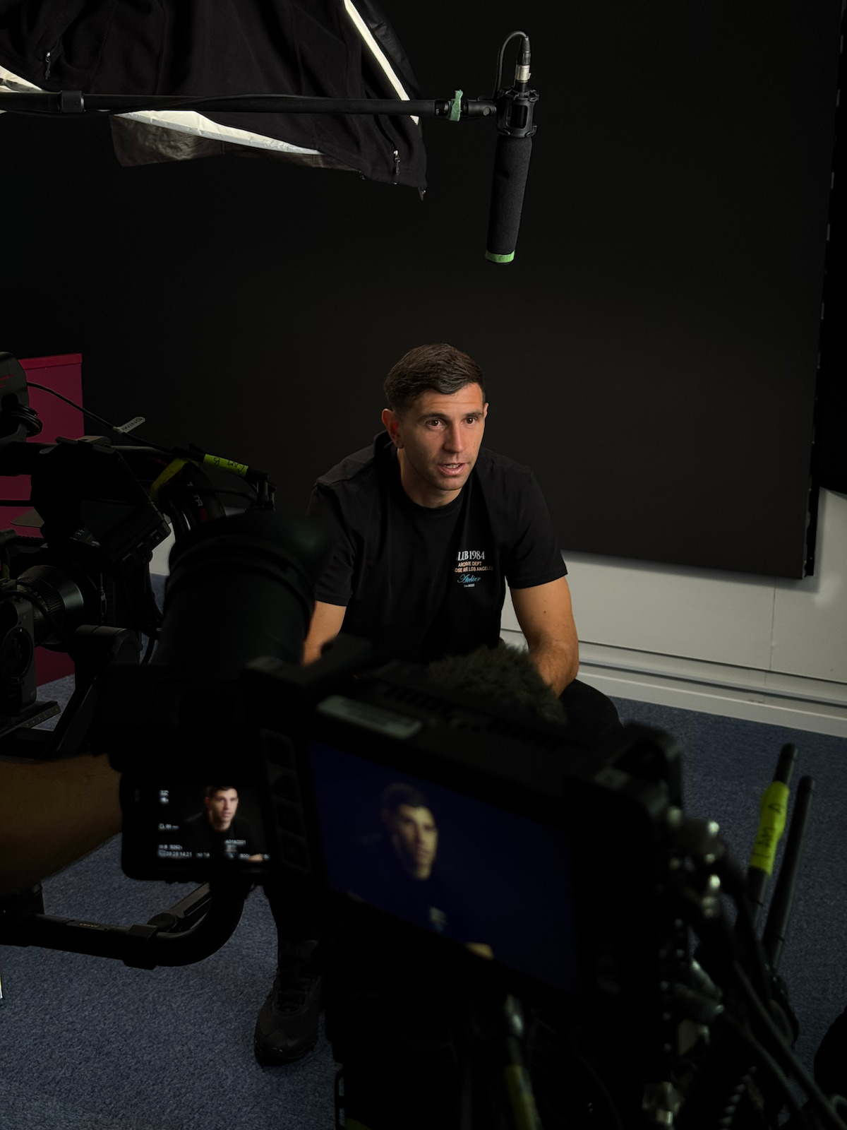 Man in black shirt sitting for a video interview in a studio, surrounded by cameras, lighting, and sound equipment, with a dark backdrop and visible recording gear.