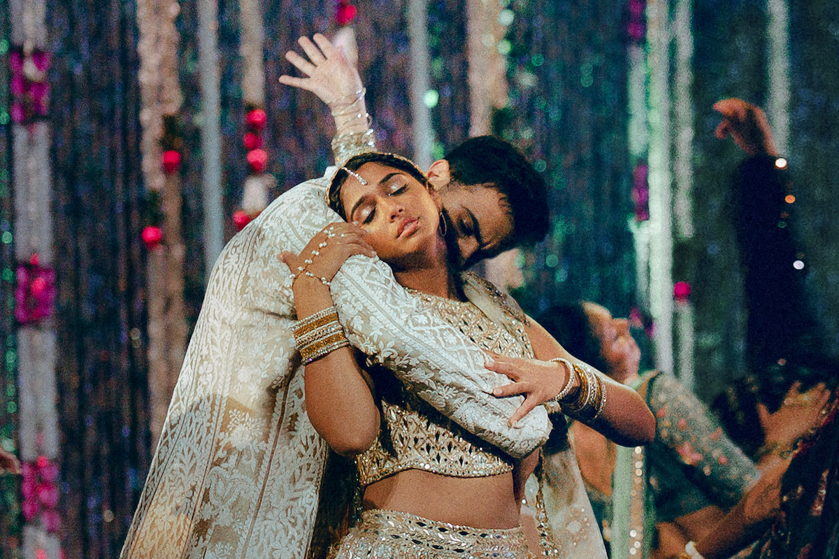 Couple in ornate traditional Indian attire dance closely at a festive event, surrounded by others in colorful clothing and shimmering streamers, creating an elegant, celebratory atmosphere.