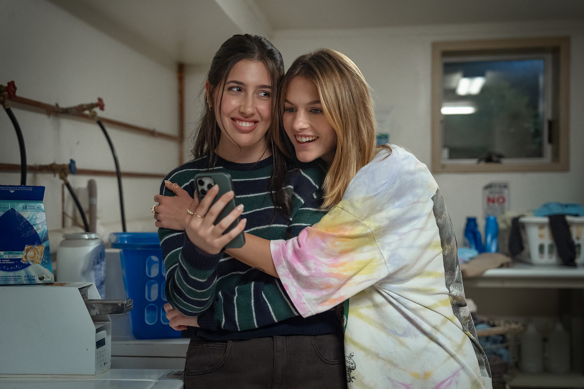 Two teenage girls smiling and taking a selfie together in a laundry room, surrounded by laundry machines and household items, with one girl hugging the other.