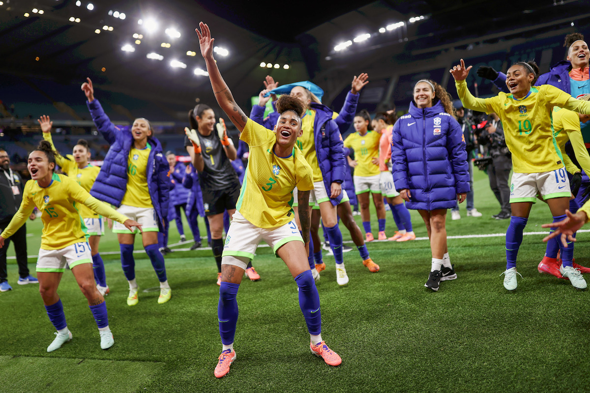 Brazilian women's soccer team celebrating on a brightly lit stadium field, players in yellow jerseys and blue shorts smiling, raising arms and dancing, with some team staff and substitutes in the background. Nighttime sporting event atmosphere.