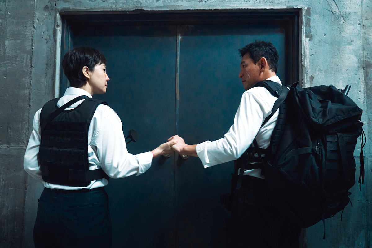 Yum Jung-ah as Kang Mi-seon and Hwang Jung-min as Park Kang-mu, both wearing black pants and white button-down shirts with tactical gear over them, hold hands in front of a closed elevator door in a frame from the movie “Mission: Cross.”