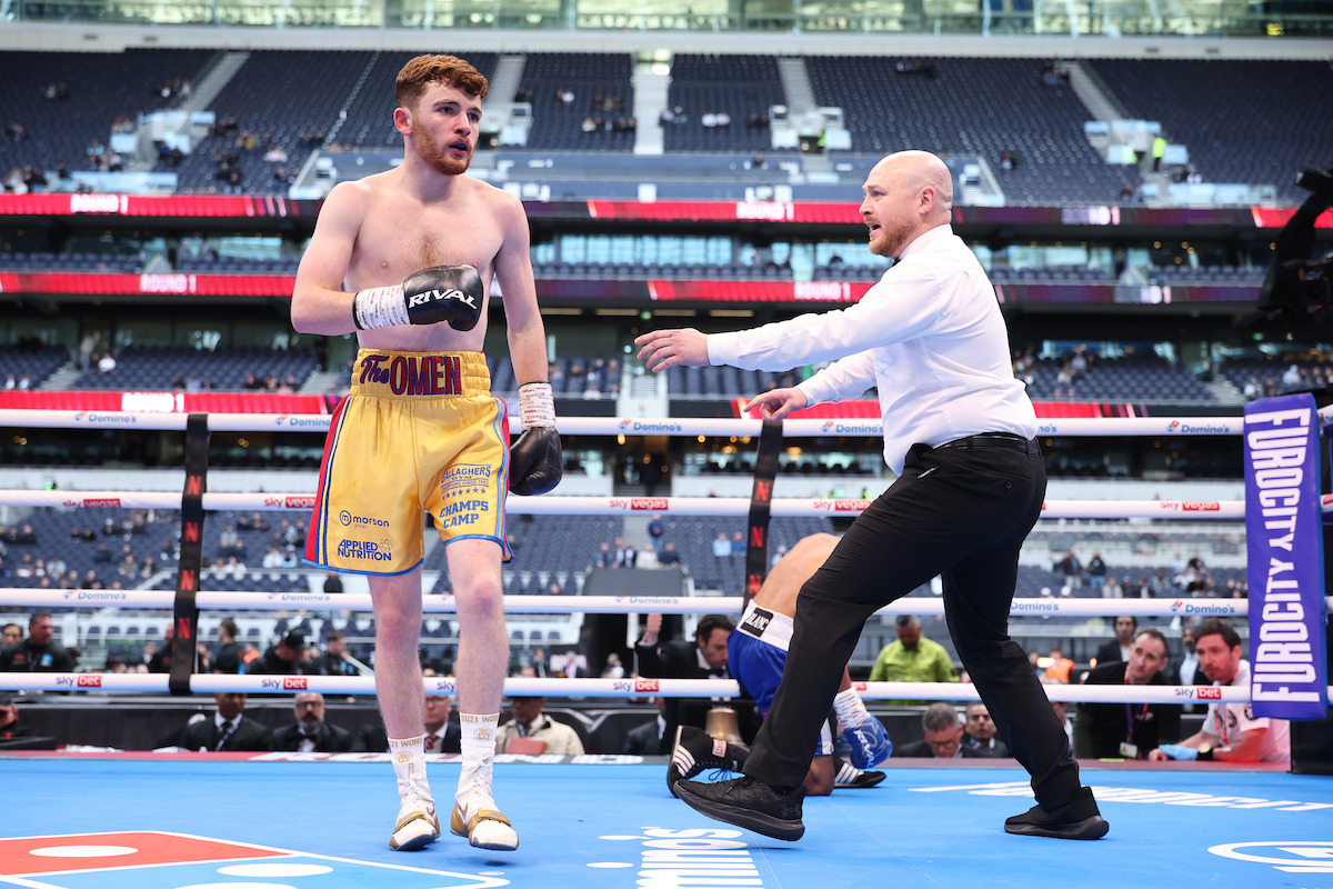 A boxer in yellow trunks stands in a ring with a referee signaling, while another boxer sits on the mat. The event takes place in a large, mostly empty stadium with rows of blue seats and bright lighting.