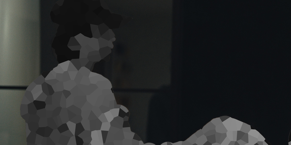 A person wearing a hat sits indoors in a dimly lit environment near a reflective surface, with much of the image pixelated and details obscured.