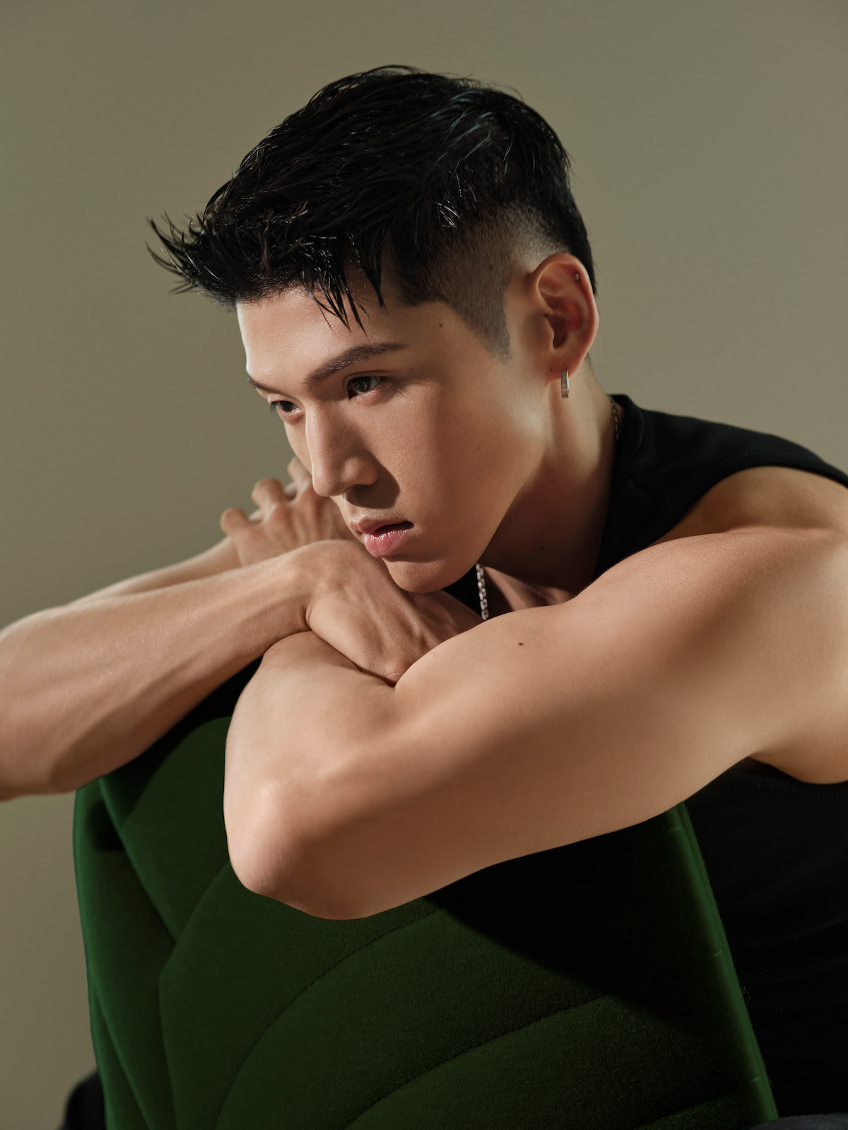 Young man with short dark hair and sleeveless shirt sits indoors, resting his arms on the back of a green chair, looking contemplative in soft, neutral lighting.