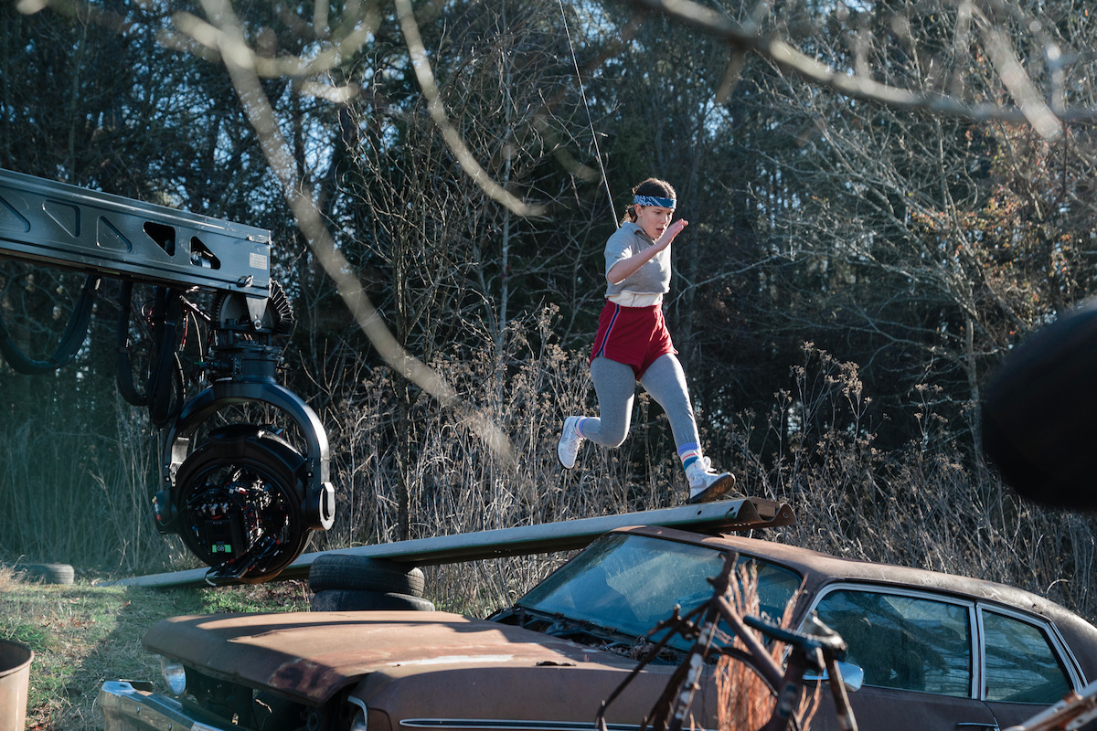 Young actor in athletic clothes sprints along a narrow plank above a rusty car as a camera rig tracks alongside.