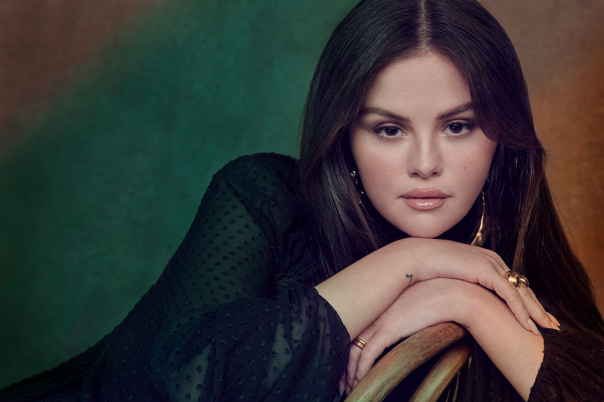 Selena Gomez wears a black top and poses on a chair against a green background.