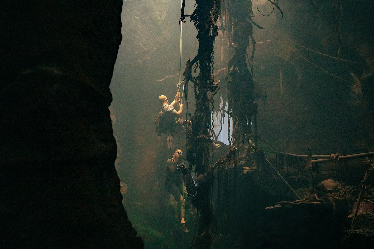 Two people climb a vine-covered rope inside a dark, misty cave with tangled vegetation and a wooden bridge, illuminated by a shaft of light from above.
