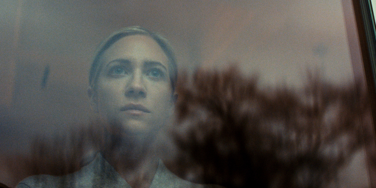 A woman with a calm, contemplative expression looks out a window, her face partially reflected in the glass with tree branches outside, evoking a quiet, introspective mood.