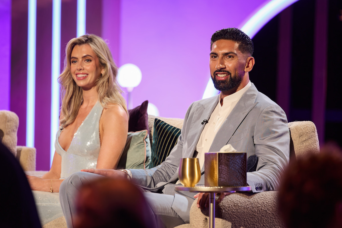 Two well-dressed people sit on a modern talk show set with purple lighting, decorative pillows, and drinks on a small table, appearing relaxed and smiling during a conversation.