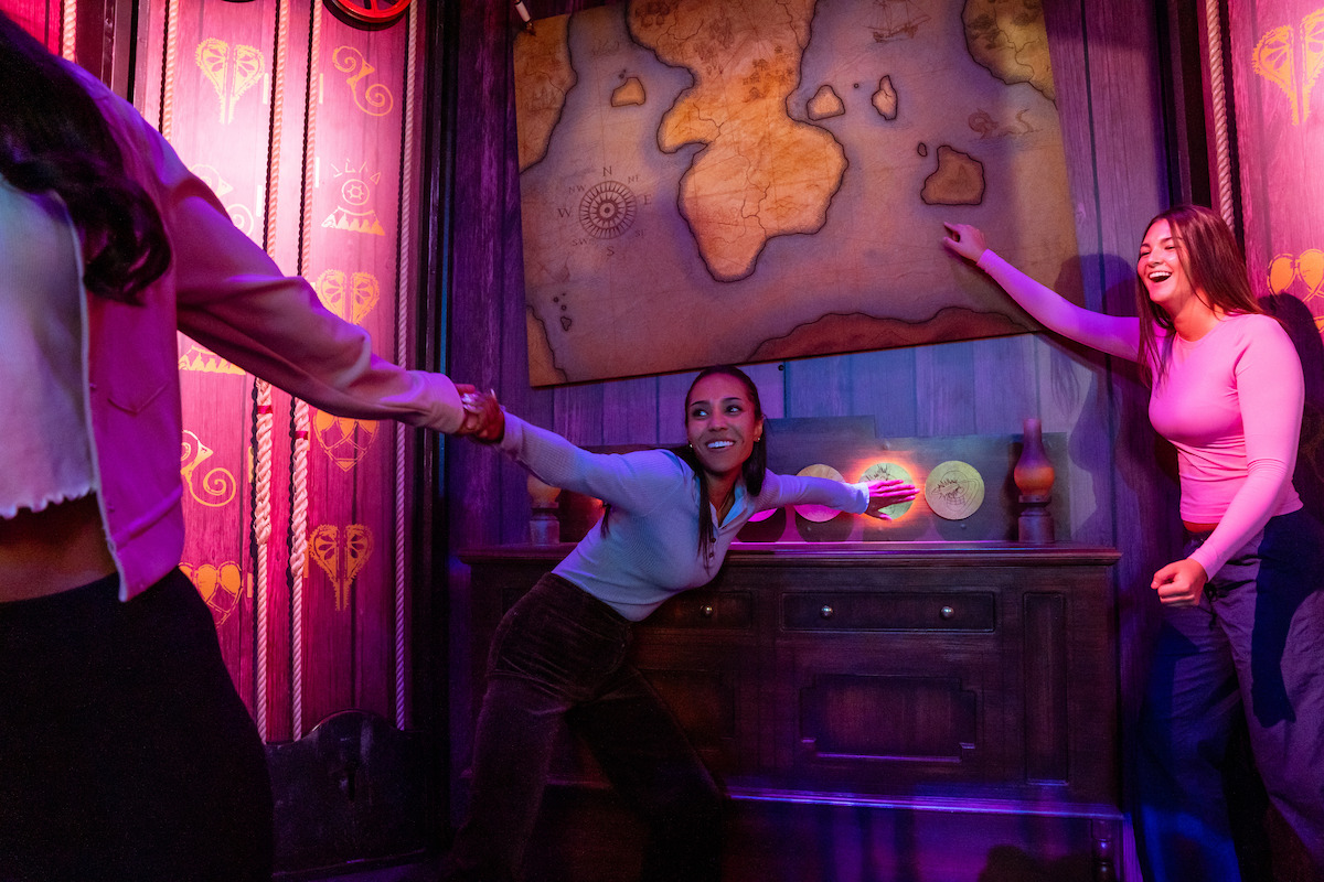 Three women enthusiastically solve a puzzle together in a colorful, themed escape room with wooden walls, glowing lights, and a large, old-style world map on the wall, creating a sense of adventure and teamwork.