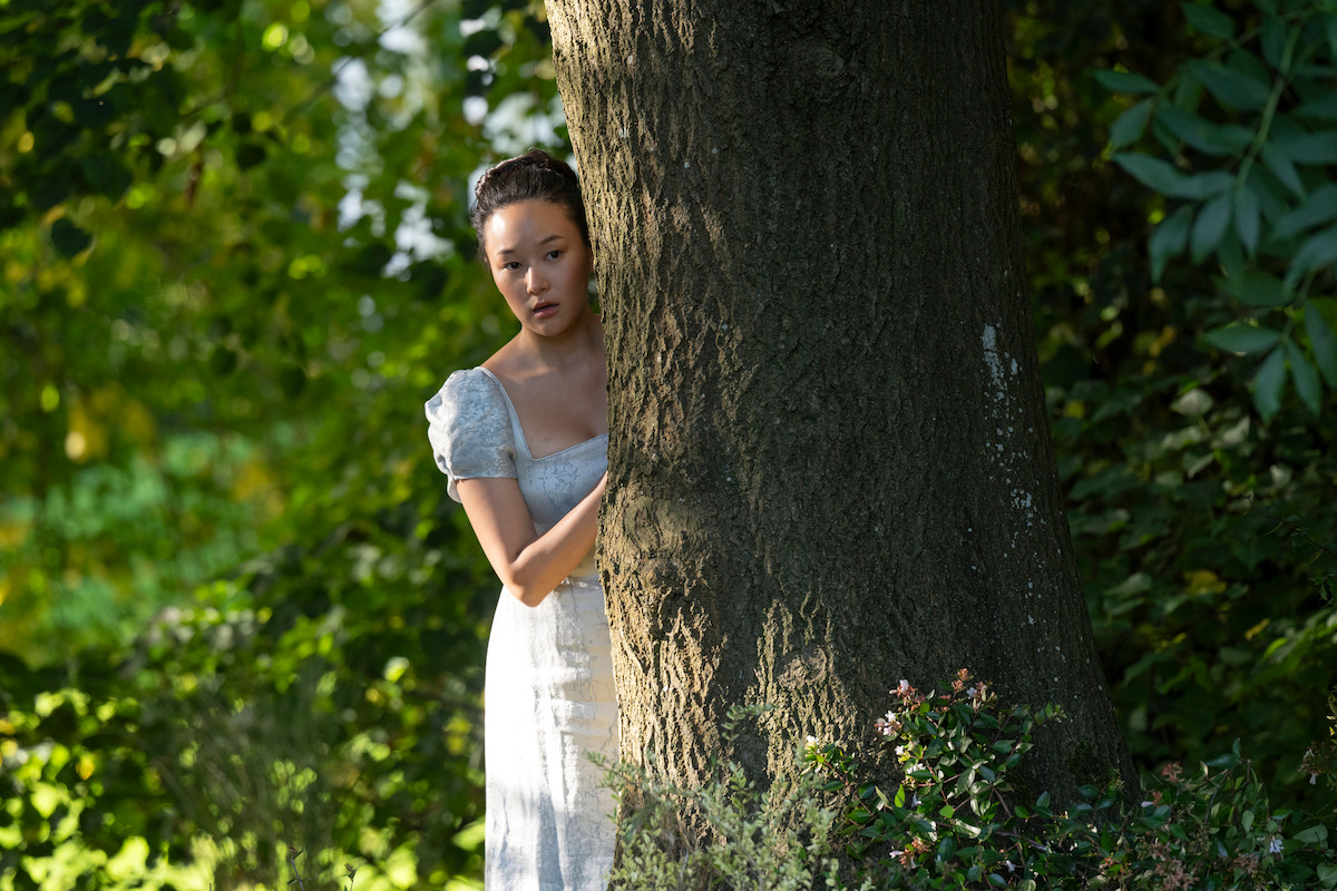 Woman in a light dress stands partially hidden behind a large tree in a sunlit, leafy forest or garden, looking out with a cautious or curious expression.