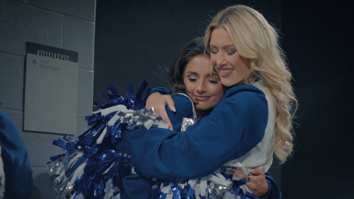 Two cheerleaders in blue and white uniforms hug backstage near a sign that says "Field Level," surrounded by blue walls. One holds pompoms as they share a close, happy moment before or after a performance.