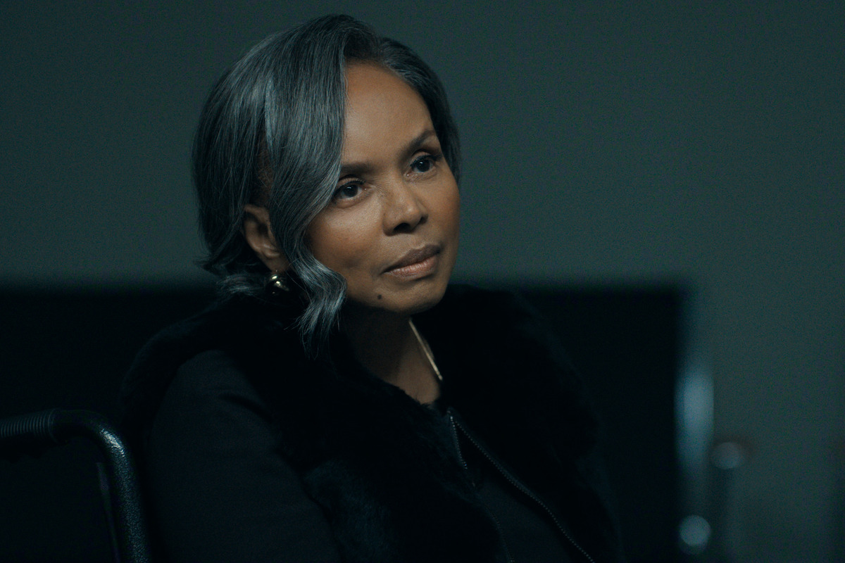 Woman with gray hair sits indoors on a black chair in a dimly lit room, wearing a dark jacket, with a neutral expression. The background is simple with minimal details, suggesting a serious or contemplative environment.