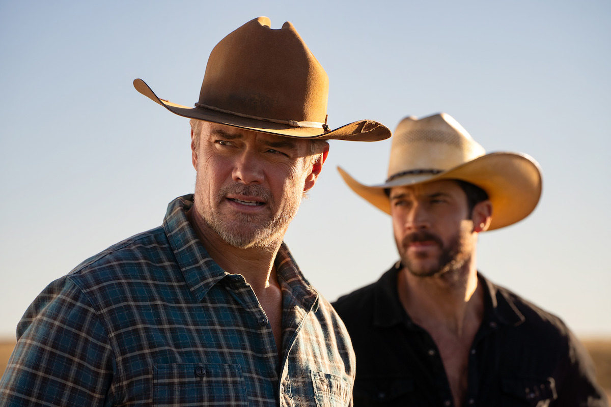 Two men wearing cowboy hats and plaid shirts stand outdoors in a rural setting with clear blue sky, sunlight highlighting their faces, evoking a western or ranch atmosphere.