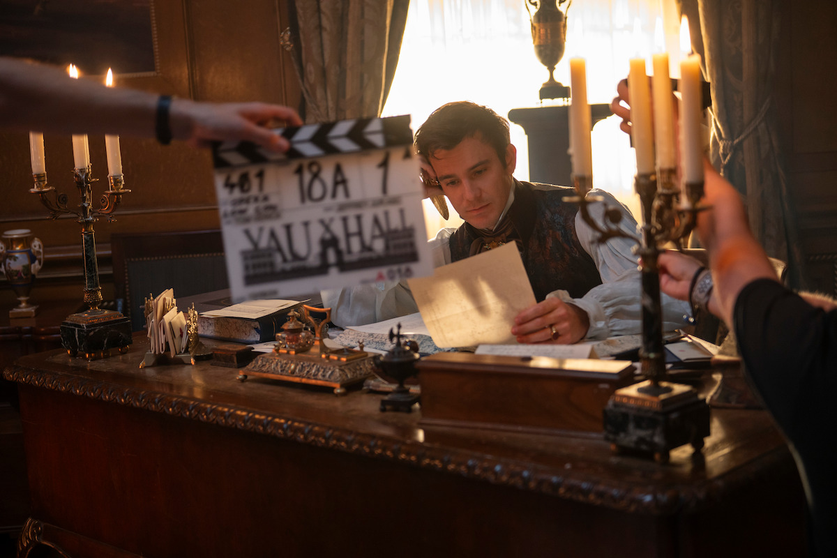 An actor in period costume sits at an ornate desk reading a letter on a vintage film set, surrounded by candles and props, as crew members hold a clapboard and adjust lighting in a warmly lit, lavishly decorated room.