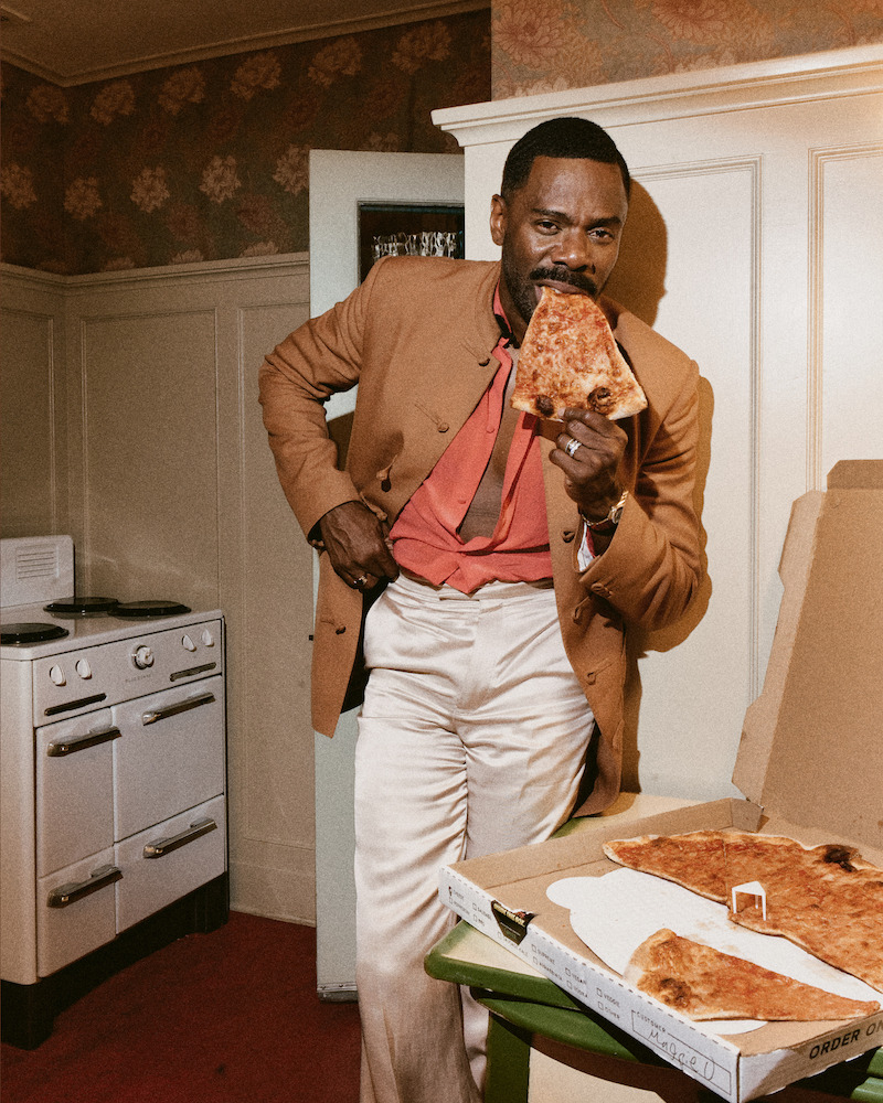 Man in a tan jacket and red shirt eating a large slice of pizza in a vintage-style kitchen with a white stove, green table, and open pizza box. Warm lighting and floral wallpaper complete the retro setting.