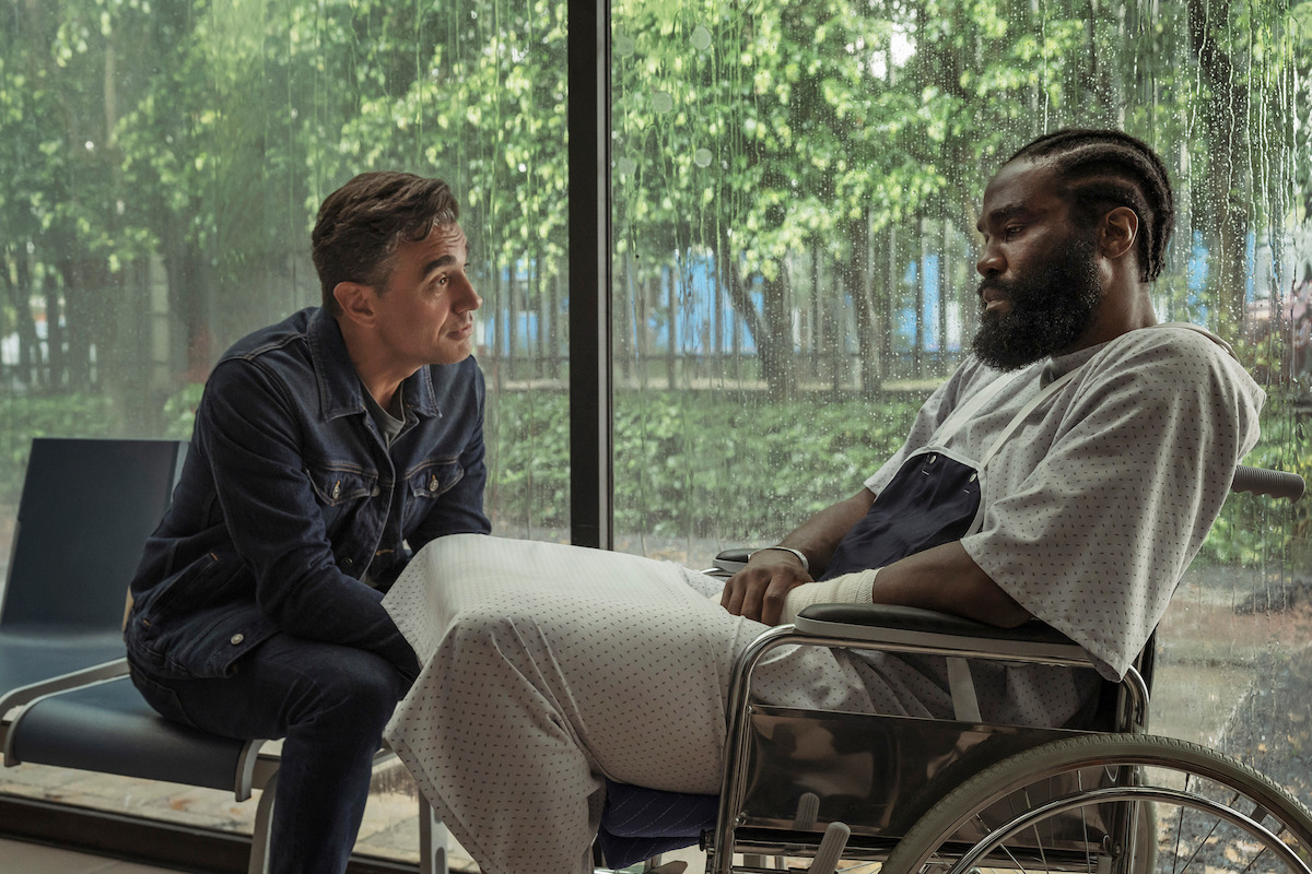 A man in a denim jacket sits and talks to another man in a hospital gown with bandaged hands, sitting in a wheelchair by a large window on a rainy day, with trees visible outside.